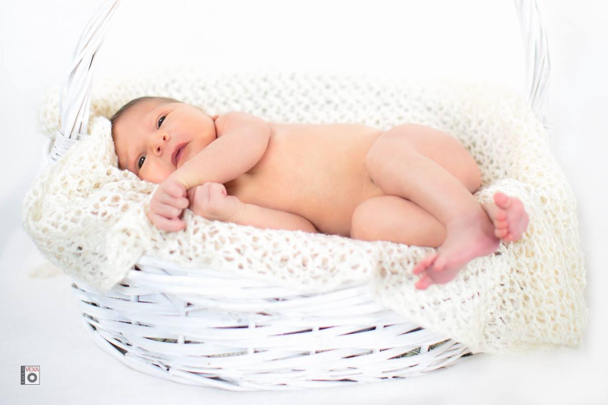 Noworodek leży na plecach w białym koszu wyłożonym kremową, ażurową tkaniną, z zaciśniętą piąstką i otwartymi oczami.