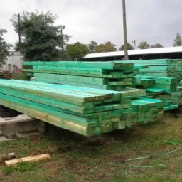 Stosy impregnowanej na zielono więźby dachowej ułożone na trawie przed magazynem, zabezpieczone taśmą, widoczne ślady po transporcie.