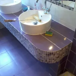 Dwie białe umywalki nablatowe na fioletowym blacie wykończonym mozaiką; na jednej z umywalek leży wałek malarski, a obok gąbka.