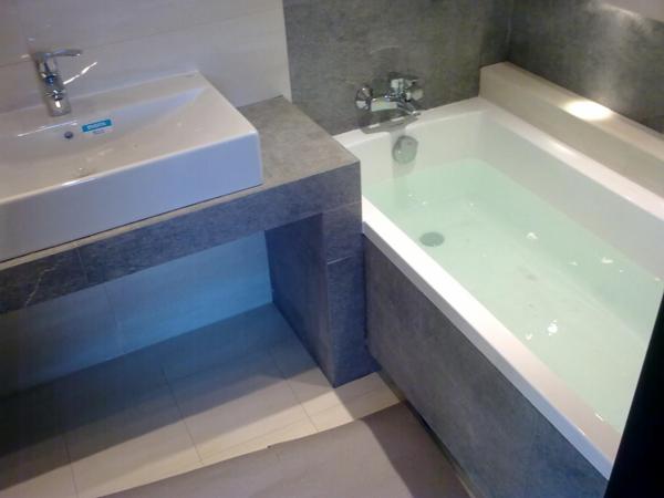 Nowoczesna łazienka z prostokątną umywalką nablatową i wanną wypełnioną wodą, wykończona szarymi płytkami imitującymi kamień.