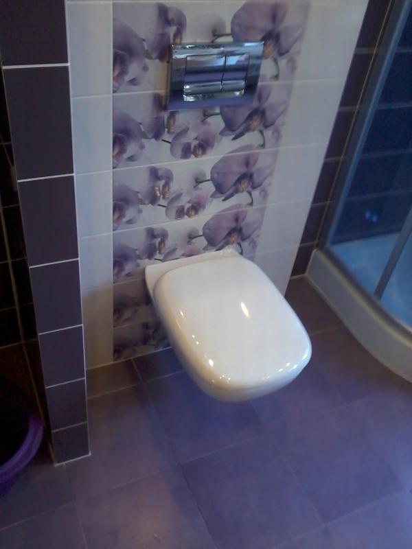 Podwieszana toaleta z chromowanym przyciskiem spłukiwania na ścianie wyłożonej płytkami z motywem fioletowych kwiatów, fioletowa podłoga z płytek.