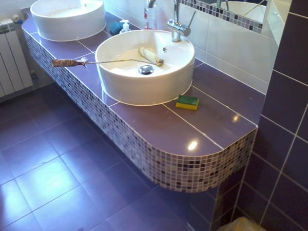 Dwie białe umywalki nablatowe na fioletowym blacie wykończonym mozaiką; na jednej z umywalek leży wałek malarski, a obok gąbka.