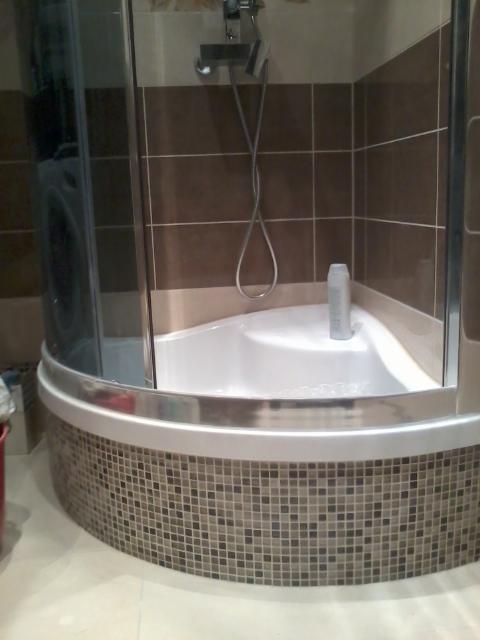 Narożna kabina prysznicowa z mozaikową obudową, brązowe płytki na ścianach i butelka szamponu na brodziku.