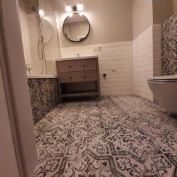 Alan-Kam - Wykończona łazienka z wzorzystą podłogą, brązową szafką pod umywalką, białą armaturą i prysznicem za szklaną ścianką.