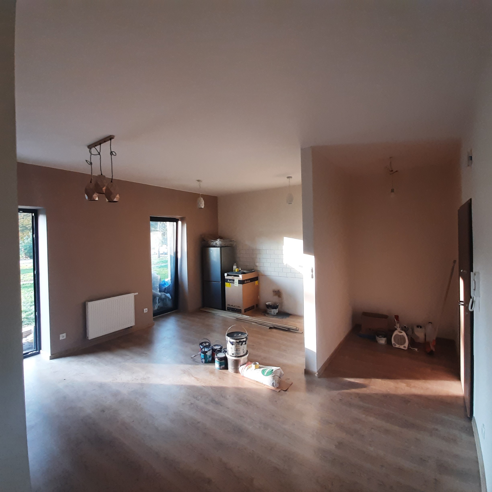 Wnętrze mieszkania w trakcie prac wykończeniowych: pomalowane ściany w odcieniach brązu, widoczne puszki z farbą na podłodze, grzejnik, okna oraz fragment kuchni z białymi płytkami.