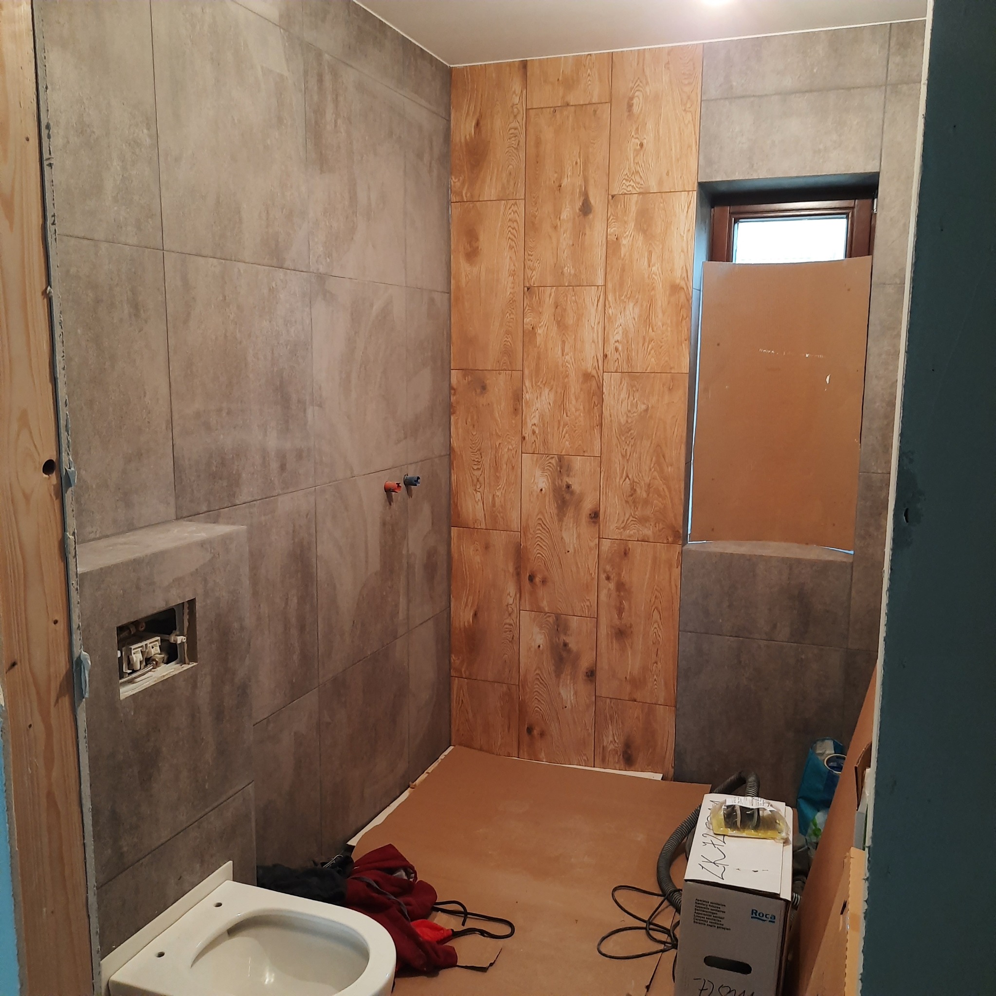 Łazienka w trakcie układania płytek: szare płytki na ścianach, drewnopodobne panele na jednej ze ścian, częściowo zamontowana toaleta, widoczne rury i przygotowane podłączenia, podłoga...