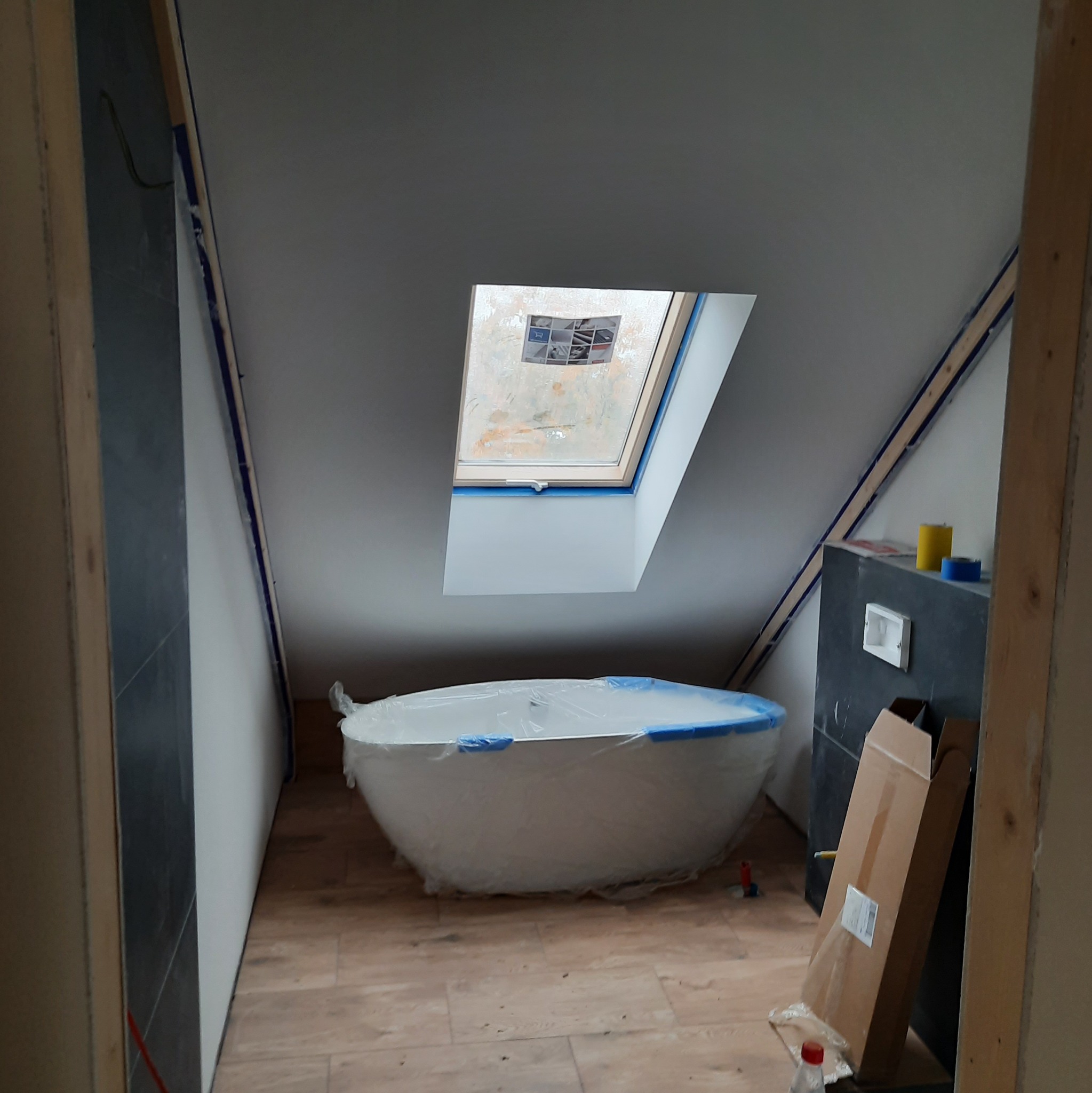 Biała wanna owinięta folią ochronną w surowym stanie łazienki na poddaszu z oknem dachowym i przygotowanymi ścianami do wykończenia.