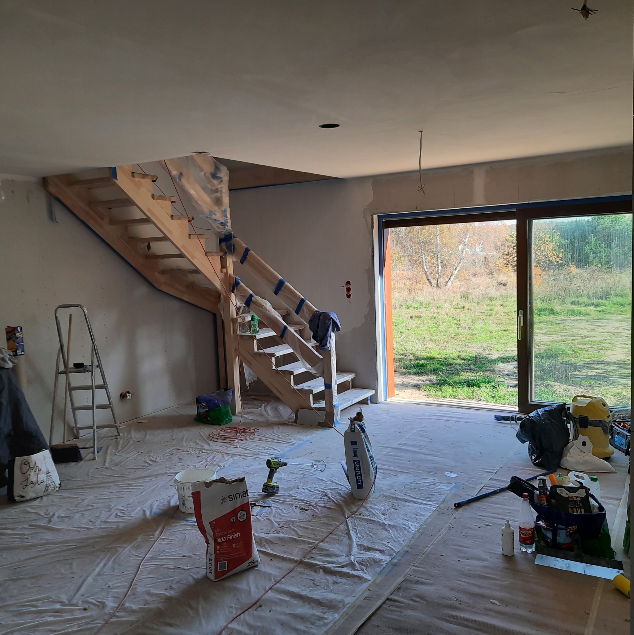 Wnętrze w trakcie prac wykończeniowych: drewniane schody zabezpieczone folią i taśmą, worki z gipsem szpachlowym, drabina oraz widok na ogród przez duże okno tarasowe.
