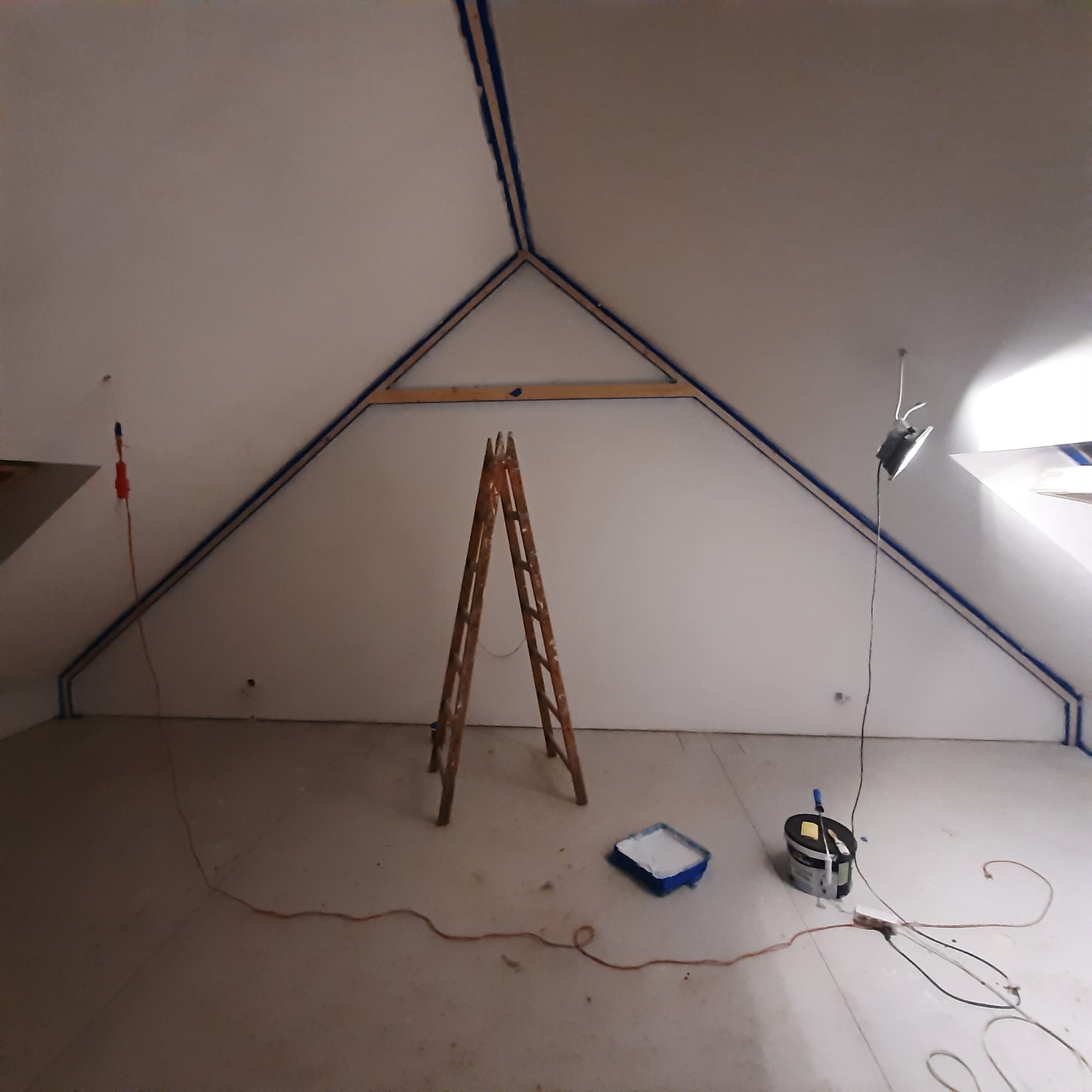 Przygotowania do malowania poddasza w Warszawie: ściany zabezpieczone taśmą malarską, drewniana drabina, kuweta z farbą i lampa oświetlająca przestrzeń.