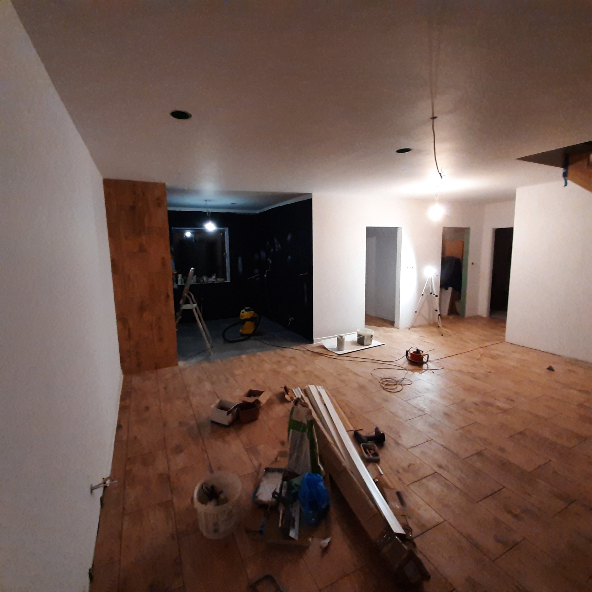 Wnętrze w trakcie prac wykończeniowych: drewniana podłoga, pomalowane ściany na biało i czarno, widoczne narzędzia, drabina i lampa na statywie.