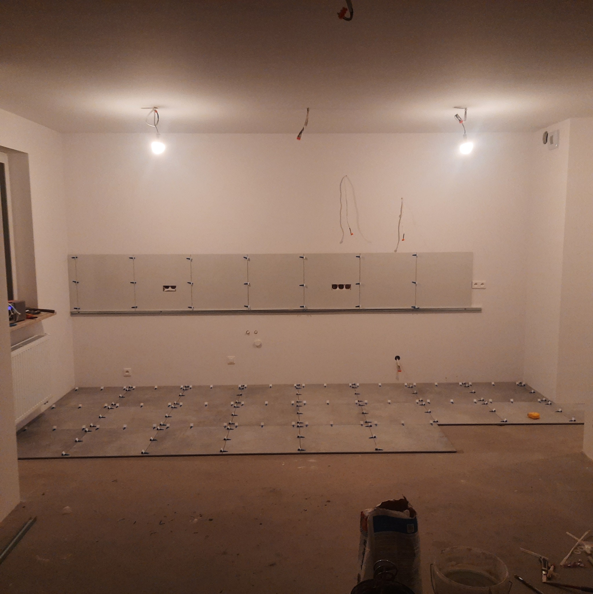 Wnętrze w trakcie układania szarych płytek podłogowych z użyciem krzyżyków dystansowych, widoczne przygotowane płytki na ścianie z zamontowanymi listwami, surowe oświetlenie i widoczne kable.
