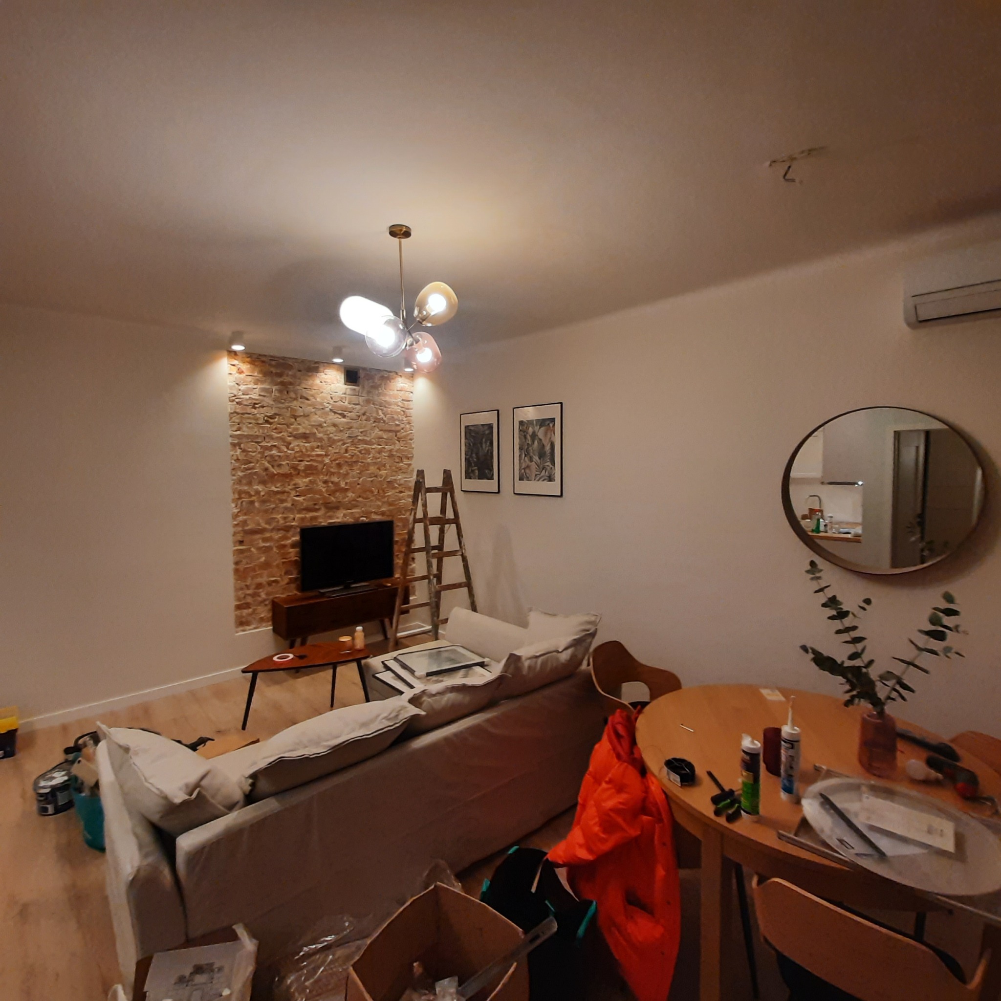 Salon w trakcie prac, widoczna ceglana ściana za telewizorem, drabina oparta o ścianę, kartony i narzędzia na podłodze, stół z silikonem i wazonem, odświeżone wnętrze.
