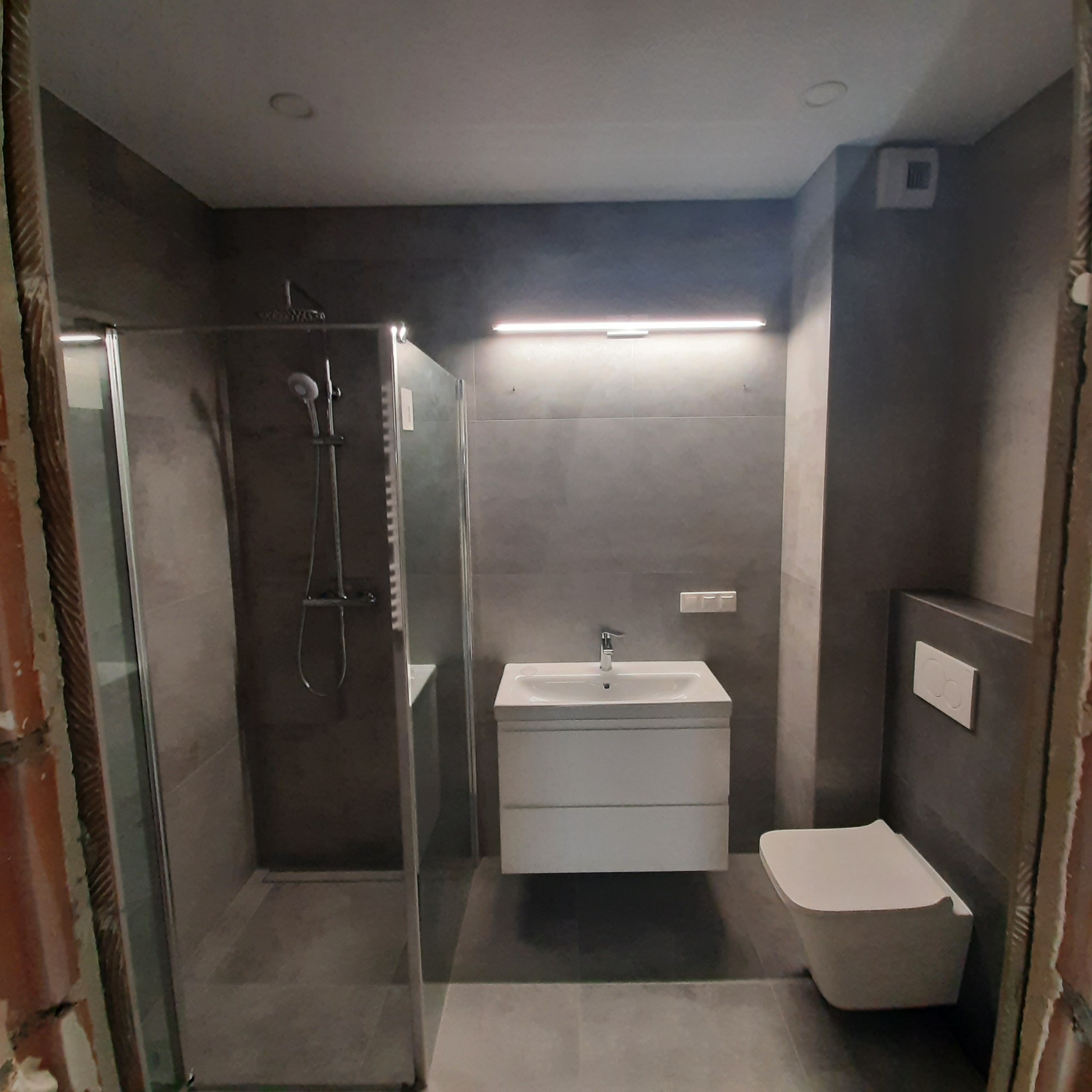 Nowoczesna łazienka z kabiną prysznicową ze szklanymi drzwiami, białą umywalką z szafką i podwieszaną toaletą, wykończona szarymi płytkami, z widocznymi surowymi brzegami ścian w trakcie prac.