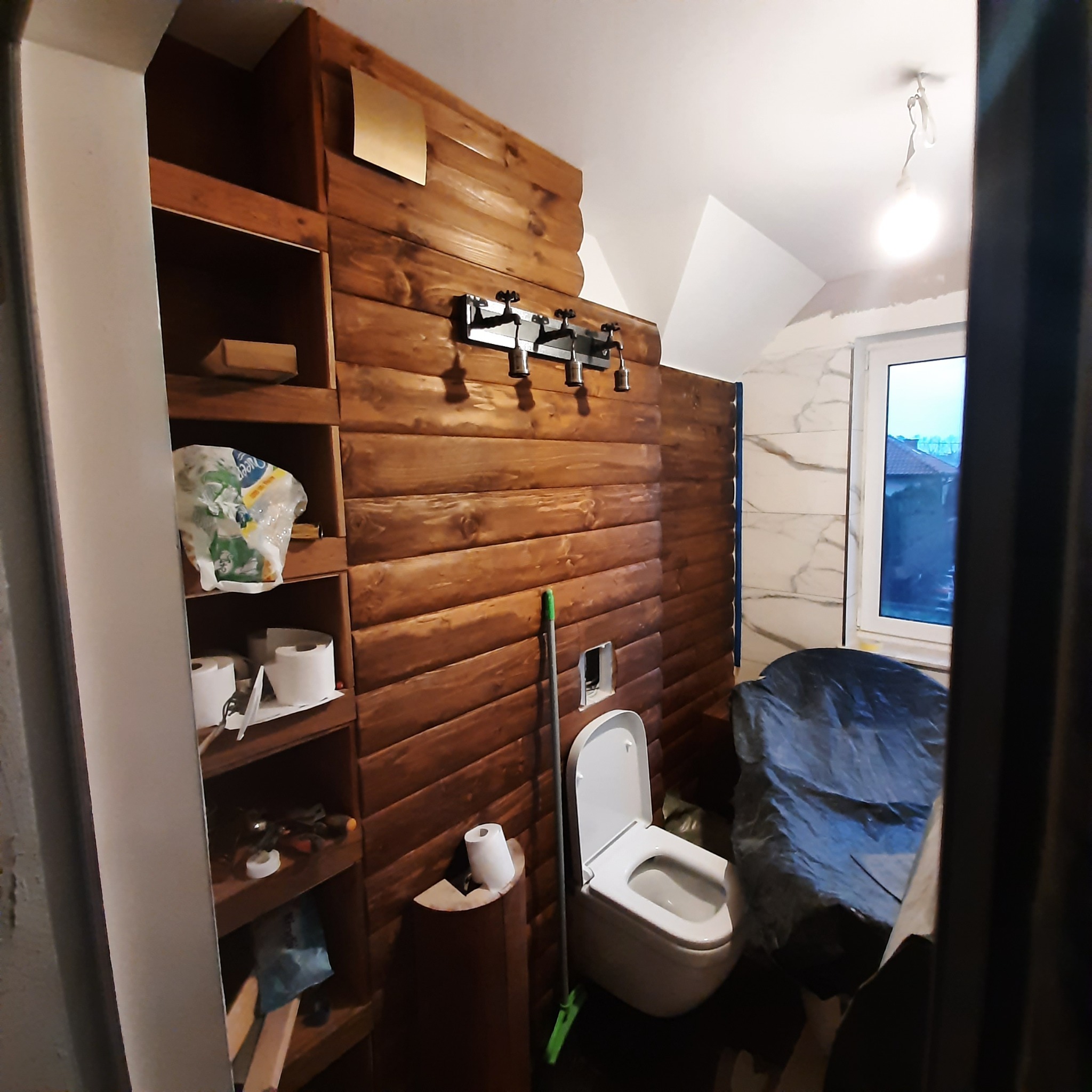 Wnętrze łazienki z drewnianą boazerią, widoczna toaleta, fragment szafki z akcesoriami i przykryte folią zabezpieczającą elementy wyposażenia podczas prac wykończeniowych, sufit częściowo...