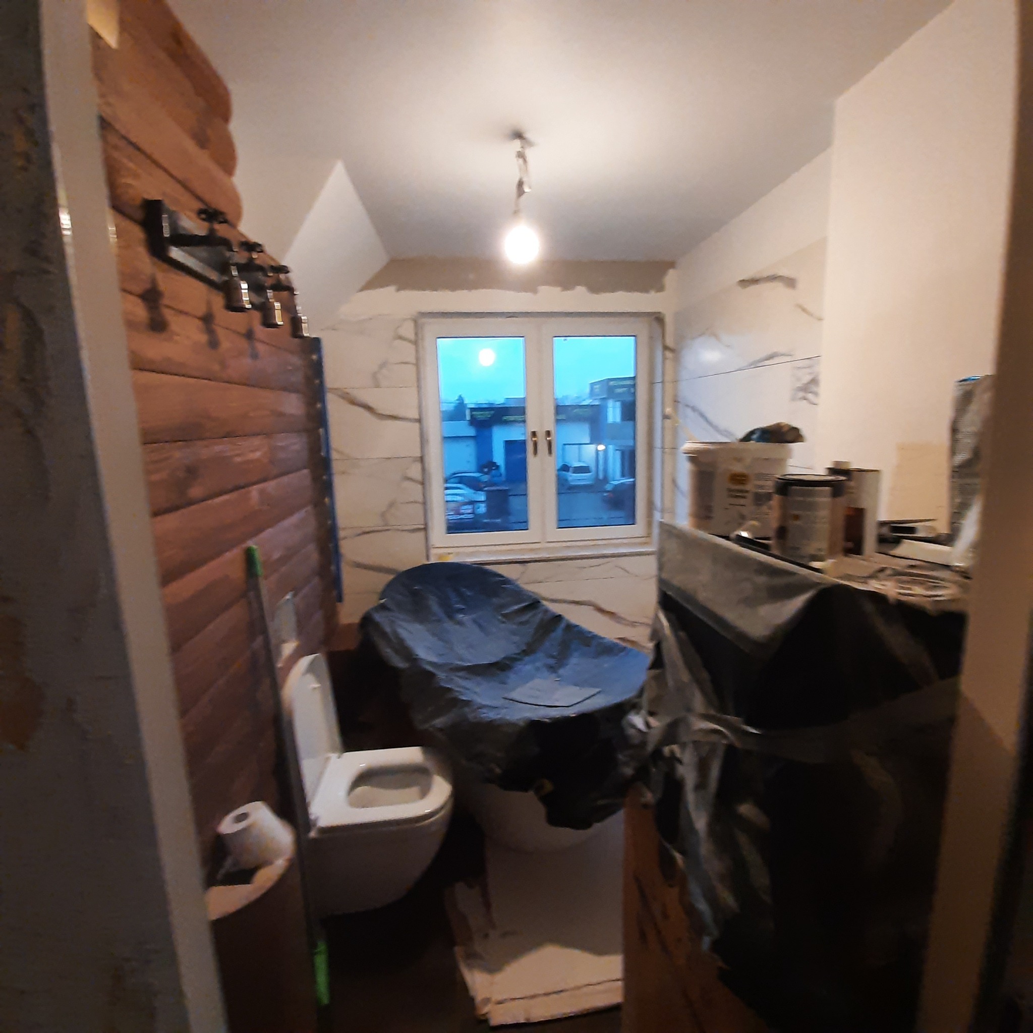 Łazienka w trakcie prac przygotowawczych do wykończenia: zabezpieczona folią wanna, przykryta toaleta, widoczne narzędzia i materiały budowlane, ściany częściowo wykończone panelami drewnianymi...