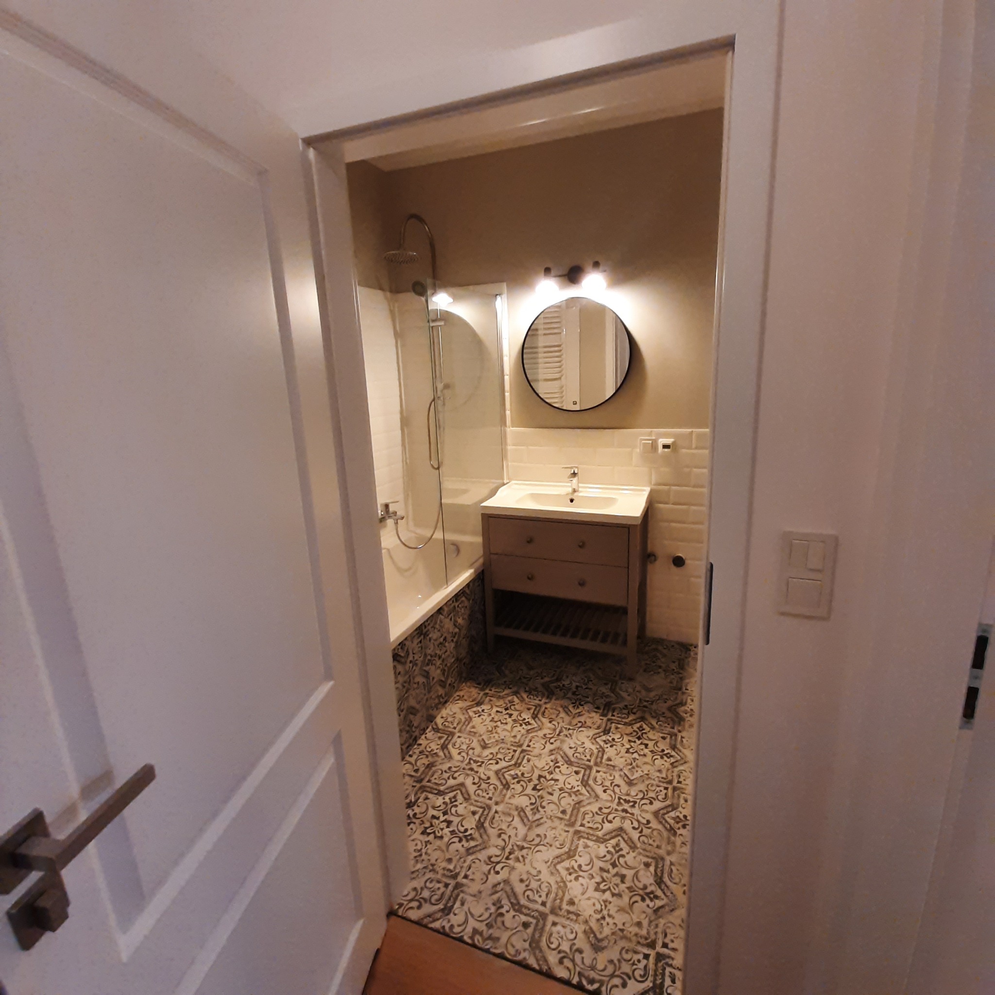 Widok na wykończoną łazienkę z kabiną prysznicową, umywalką z szafką i okrągłym lustrem, przez otwarte białe drzwi z ozdobnymi panelami i mosiężną klamką. Na podłodze wzorzyste płytki.