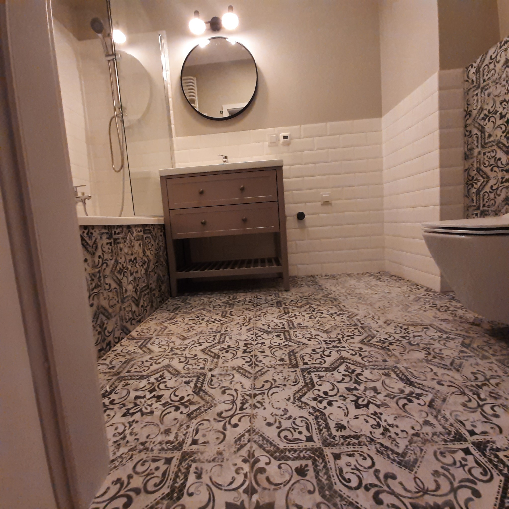 Wykończona łazienka z wzorzystą podłogą, brązową szafką pod umywalką, białą armaturą i prysznicem za szklaną ścianką.