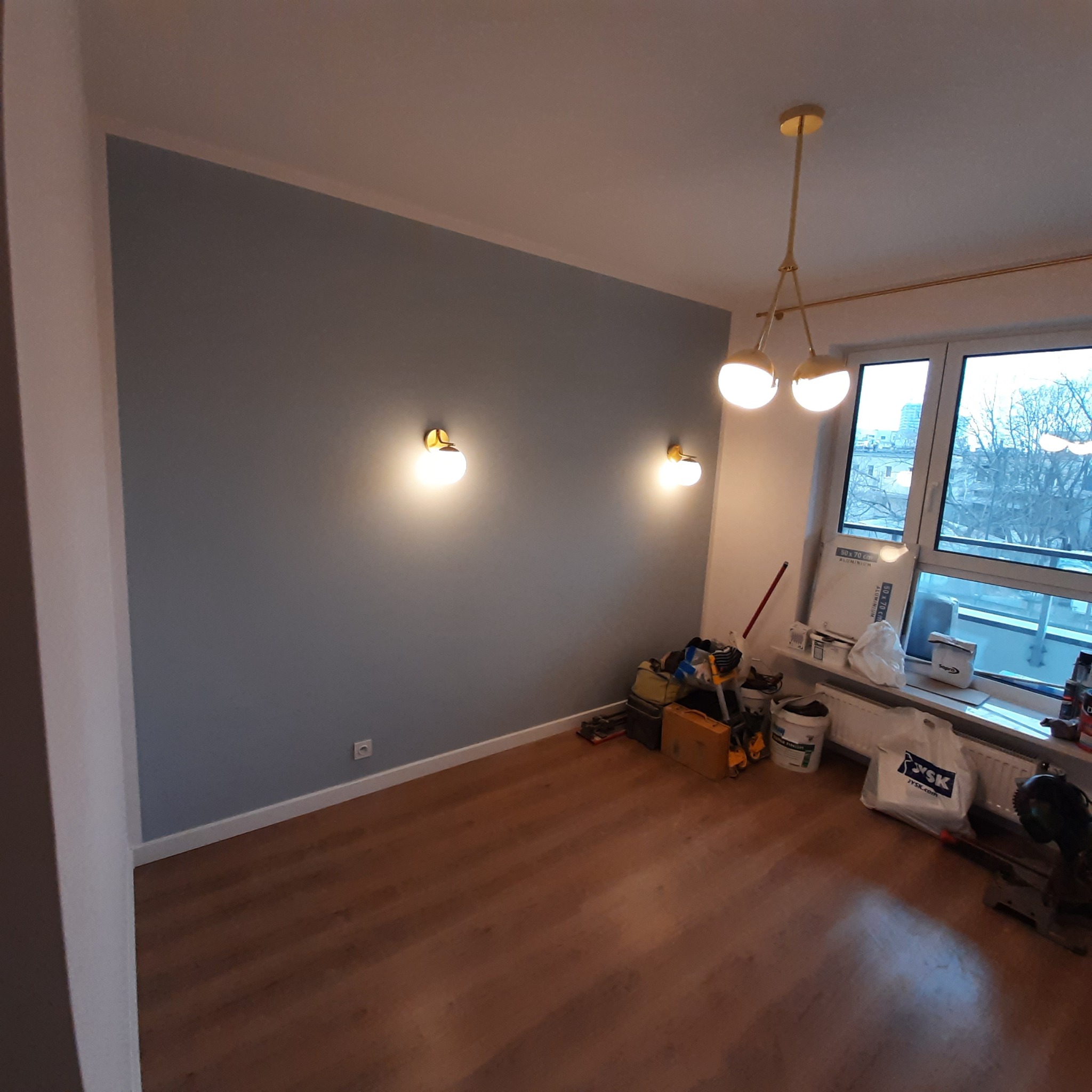 Pokój po malowaniu ścian na szaro, z widocznymi narzędziami i materiałami malarskimi przy oknie, drewniana podłoga, złote lampy ścienne i sufitowa.