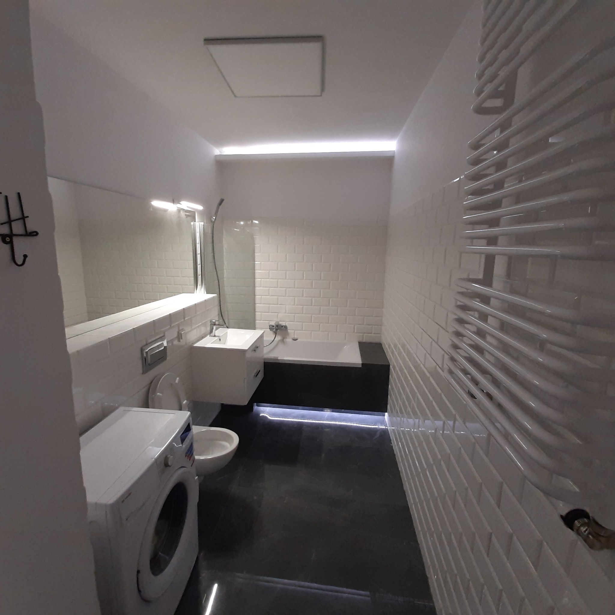 Nowoczesna łazienka w odcieniach bieli i czerni z podświetleniem LED pod wanną, widoczna pralka, umywalka, lustro i grzejnik drabinkowy.