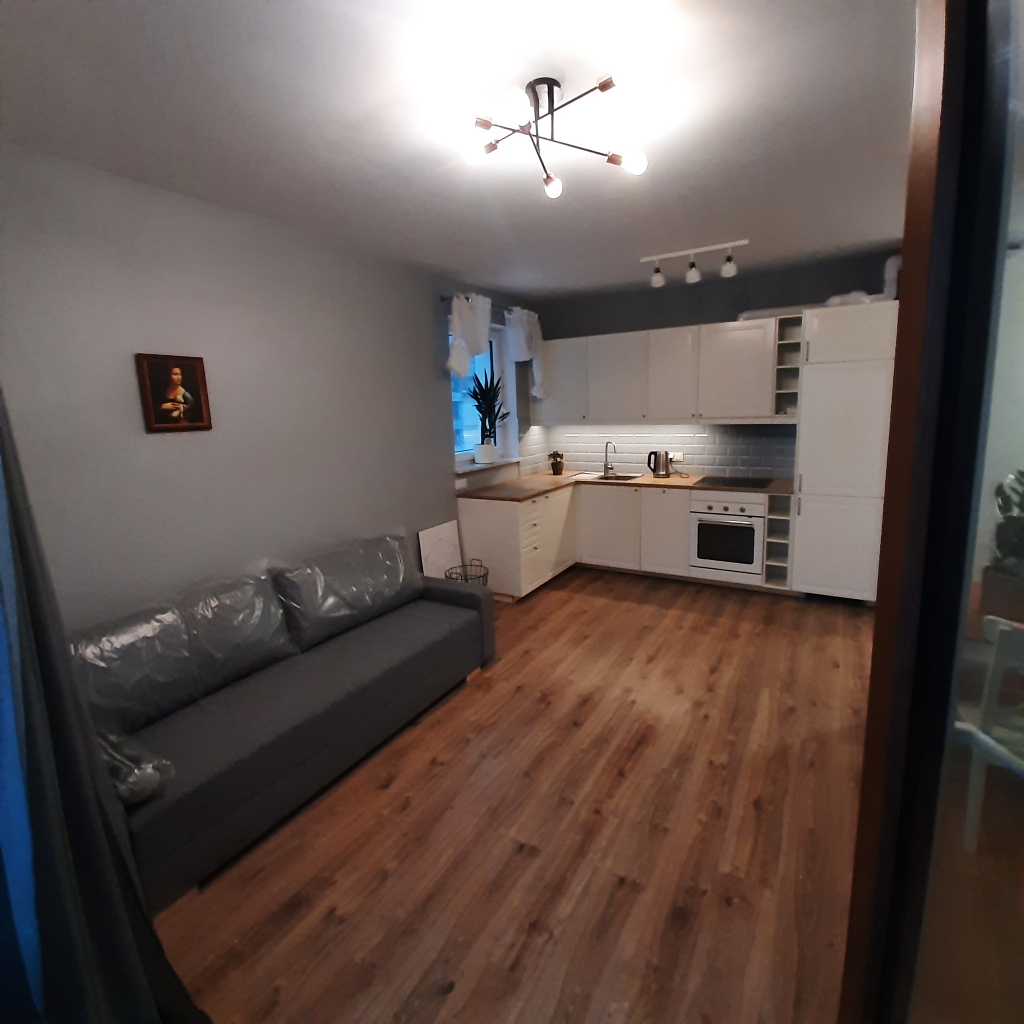 Widok na pokój z aneksem kuchennym: szara sofa z folią ochronną, parkiet, biała zabudowa kuchenna z mozaiką nad blatem.