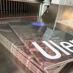 Stos zszytych broszur z okładką o teksturze kamienia pod gilotyną introligatorską, widoczny laserowy wskaźnik cięcia.