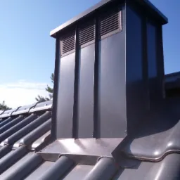 Szary, metalowy komin wentylacyjny z kratkami, zamontowany na ciemnoszarym dachu z dachówki ceramicznej, na tle błękitnego nieba.