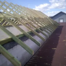 Konstrukcja drewniana dachu z belek w kolorze jasnej zieleni, widok z g&oacute;ry na częściowo pokryty dach z blachy trapezowej w kolorze ciemnego brązu, w tle fragment budynku mieszkalnego i błękitne...