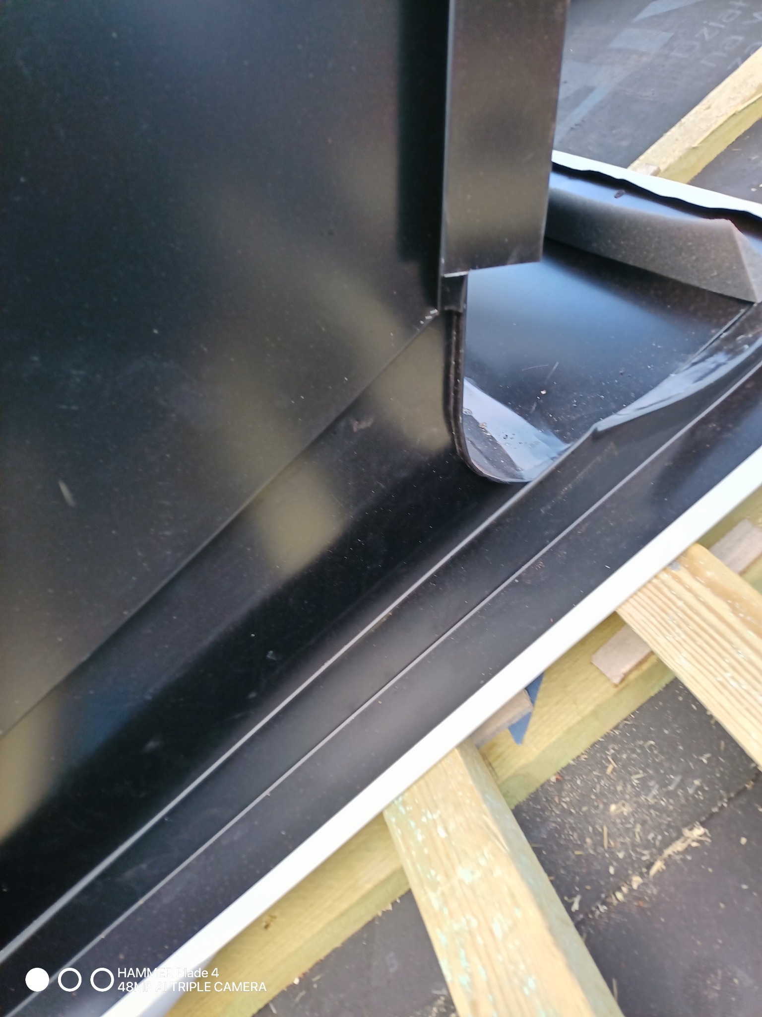 Czarny element obróbki blacharskiej, prawdopodobnie okap, zamontowany na dachu, z widocznymi elementami drewnianej konstrukcji wsporczej.