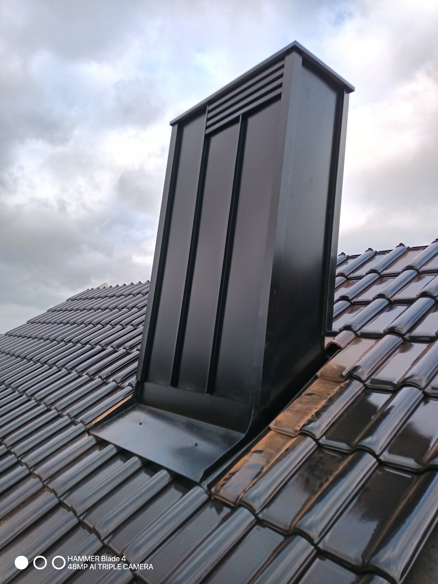 Błyszczące, grafitowe dachówki ceramiczne z kominem wentylacyjnym w kolorze. Kadr z dołu, pochmurne niebo w tle.