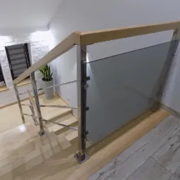 Szklana balustrada z drewnianym pochwytem i metalowymi słupkami, zamontowana na drewnianych schodach w jasnym wnętrzu z oknem i dekoracją 'HOME'.