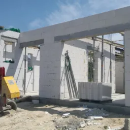 Wznoszenie ścian parterowego domu z betonu komórkowego, widoczna czerwono-żółta betoniarka, podpory i paleta z bloczkami na placu budowy w słoneczny dzień.