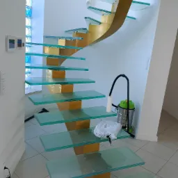 Nowoczesne schody ze szklanymi stopniami i złotą konstrukcją nośną w domu w Ołoboku. Widoczny odkurzacz obok. Nietypowe rozwiązanie architektoniczne.