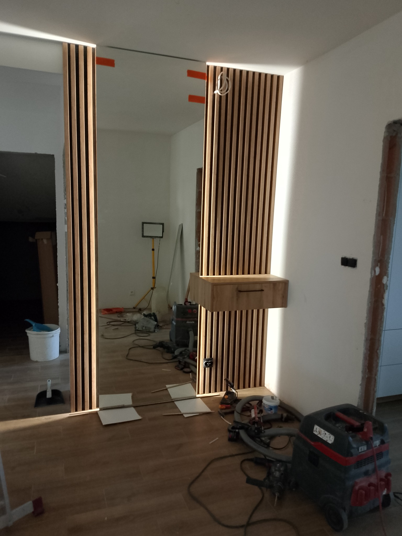 Lamelowa ściana z lustrem i drewnianą szafką w przedpokoju, widoczne narzędzia i odkurzacz. Montaż w trakcie prac wykończeniowych.