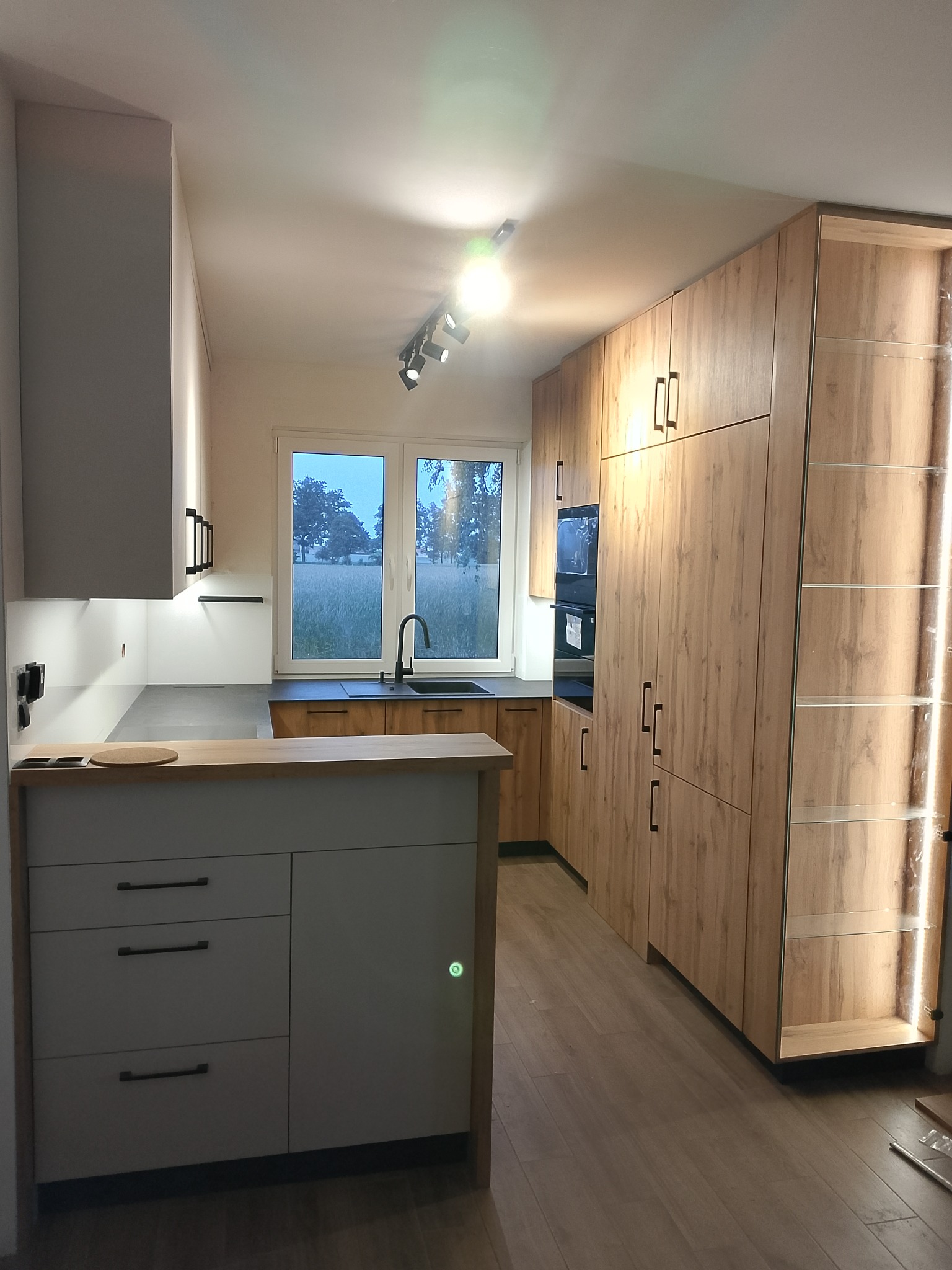 Nowoczesna kuchnia z drewnianymi szafkami i szarym blatem, oświetlona punktowo, z widokiem na zielone pole za oknem. Minimalistyczny design i funkcjonalność.