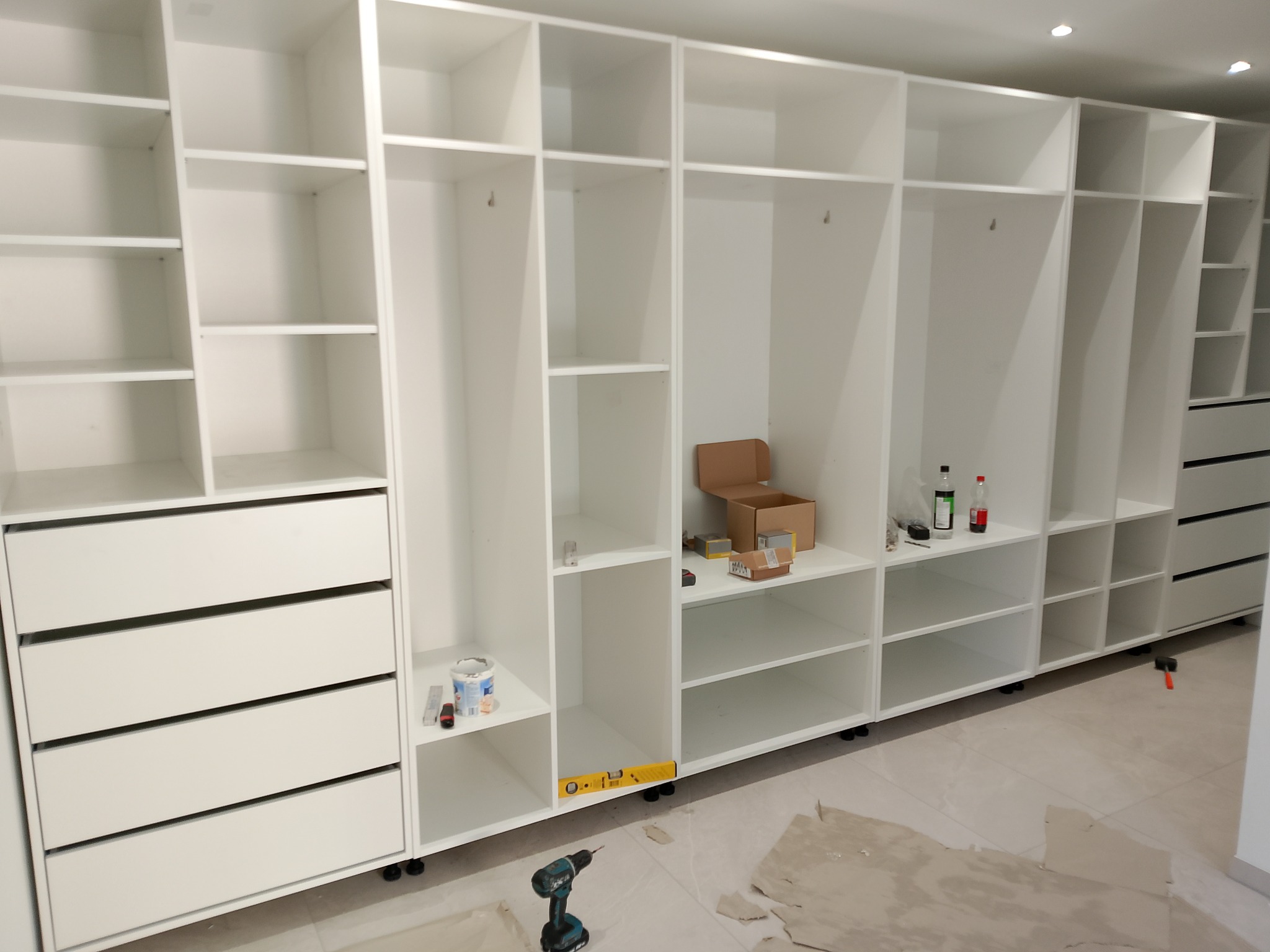 Biała szafa na wymiar w trakcie montażu, widoczne półki, szuflady, poziomica i wkrętarka. Na podłodze resztki po montażu.