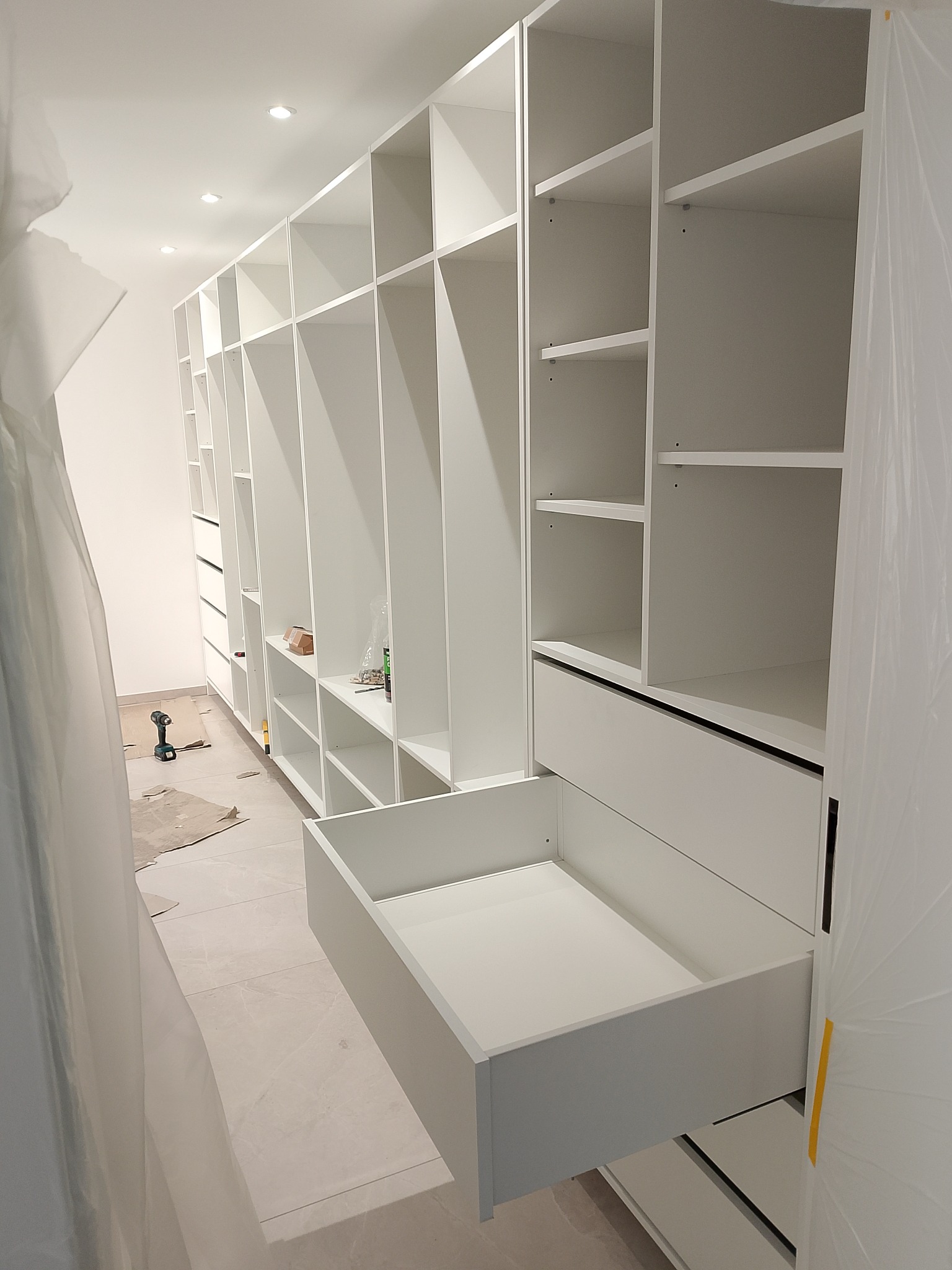 Biała, modułowa szafa na wymiar w trakcie montażu. Widoczna otwarta szuflada, półki i narzędzia. Minimalistyczny design, jasne wnętrze.