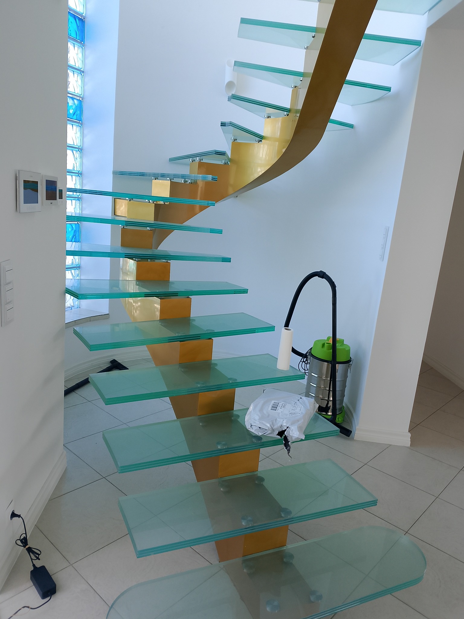 Nowoczesne schody ze szklanymi stopniami i złotą konstrukcją nośną w domu w Ołoboku. Widoczny odkurzacz obok. Nietypowe rozwiązanie architektoniczne.