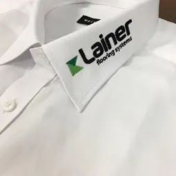 Biała koszula z haftowanym logo 'Lainer flooring systems' na kołnierzyku, widoczny guzik i metka.