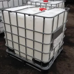 Paleta z kontenerem IBC z białą zawartością i czerwonym korkiem, otoczona metalową klatką, na nierównym, ziemistym podłożu.