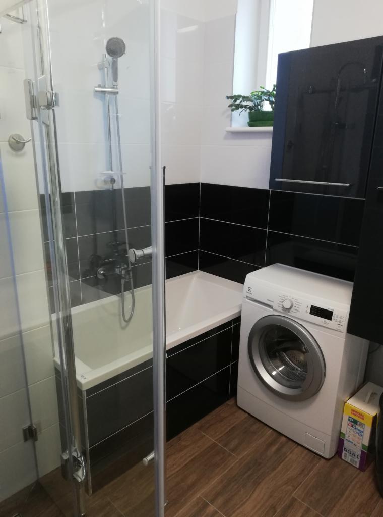 Nowoczesna łazienka z kabiną prysznicową, wanną, pralką Electrolux i czarno-białymi płytkami, drewnopodobna podłoga, roślina doniczkowa na parapecie.