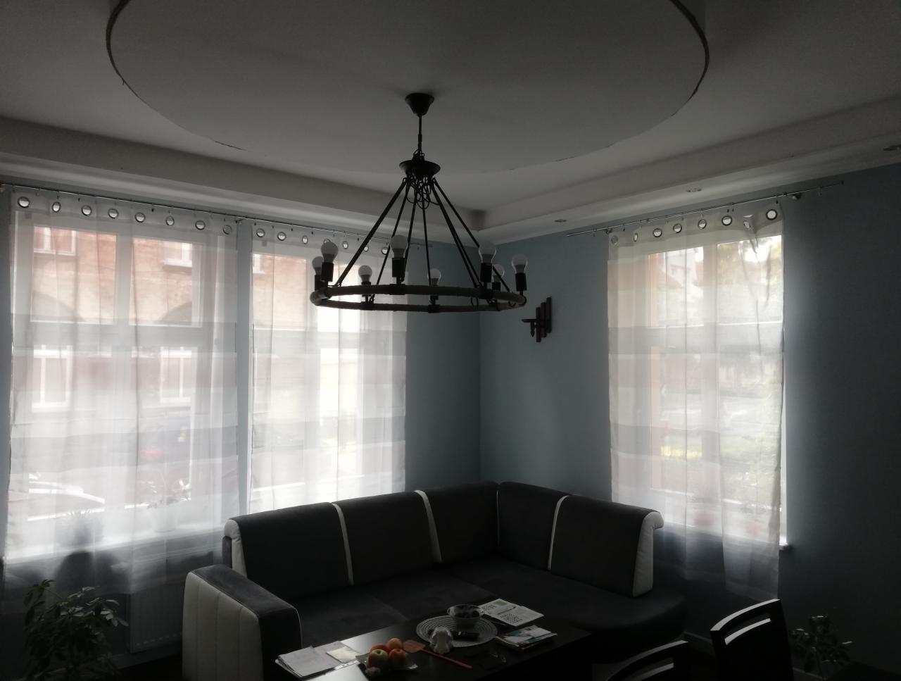 Salon z szarą sofą narożną, białymi, półprzezroczystymi zasłonami okiennymi i oryginalnym żyrandolem w stylu retro z żarówkami Edison