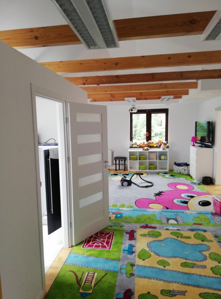 Pokój dziecięcy po sprzątaniu: widoczny kolorowy dywan z motywami dziecięcymi, białe ściany, drewniane belki na suficie i otwarte drzwi z białymi panelami.