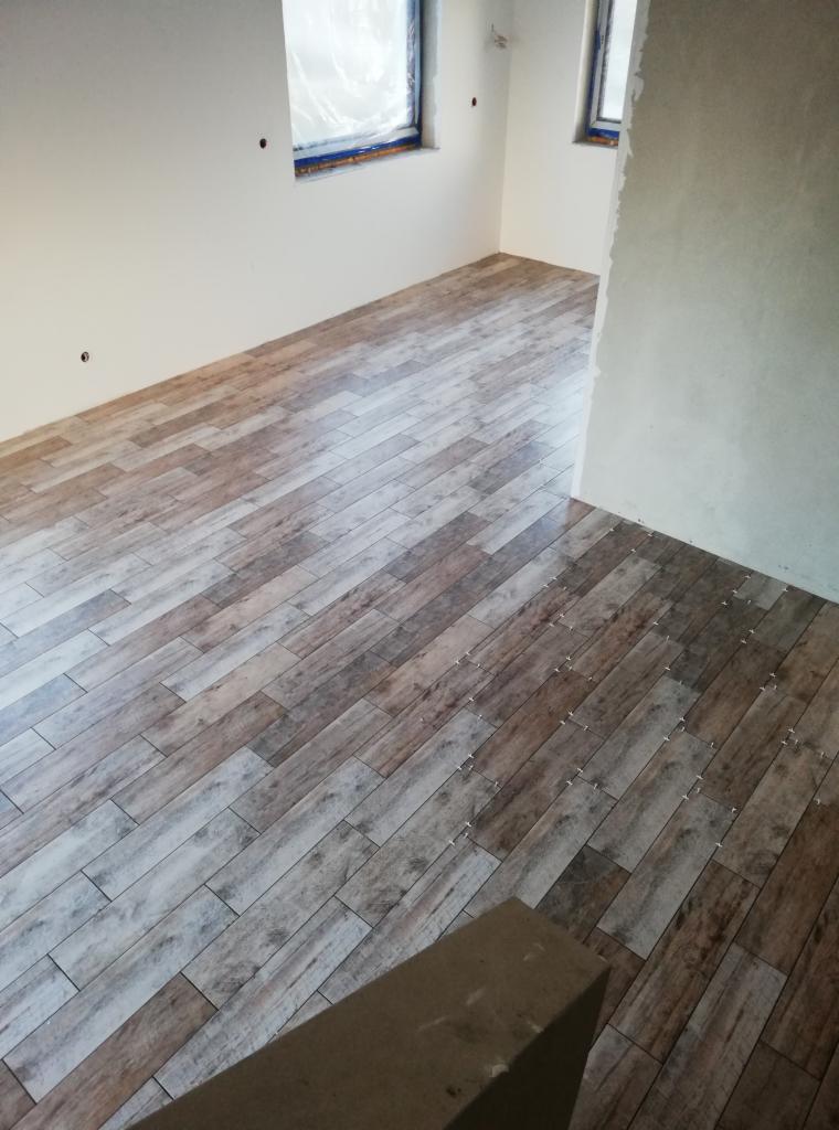 Ułożona podłoga z płytek imitujących drewno w nowym pomieszczeniu, widoczne krzyżyki dystansowe, okna zabezpieczone folią malarską.