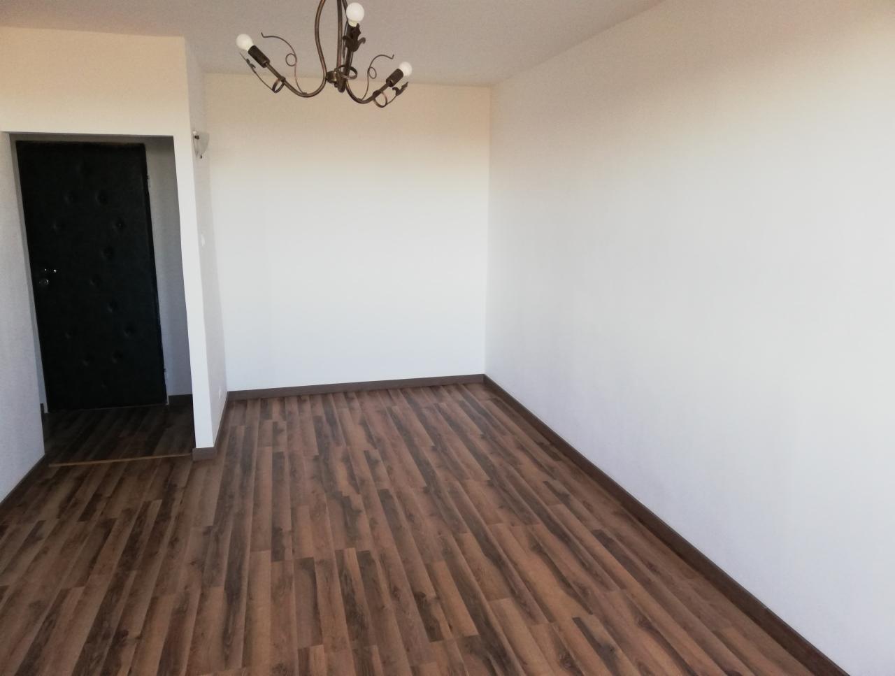 Puste pomieszczenie z panelami podłogowymi imitującymi drewno, żyrandol z kilkoma żarówkami i czarne drzwi w lewym rogu.