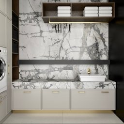Inspiration Studio - Nowoczesna pralnia z marmurową ścianą, pralką i suszarką Beko, szafkami w kolorze beżowym i złotymi akcentami. Minimalistyczny design z półkami na ręczniki.