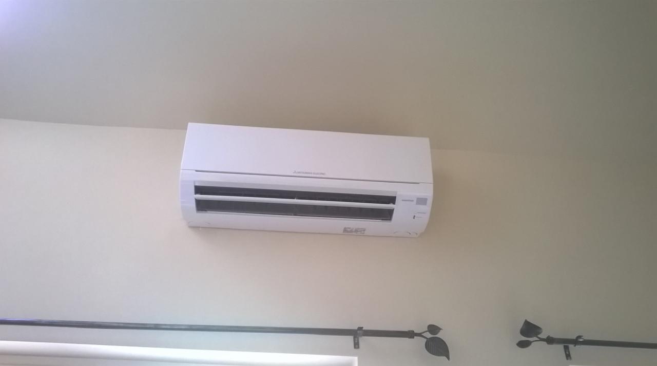 Biały klimatyzator Mitsubishi Electric zamontowany na ścianie w pomieszczeniu z beżowymi ścianami i karniszem z metalowymi liśćmi.