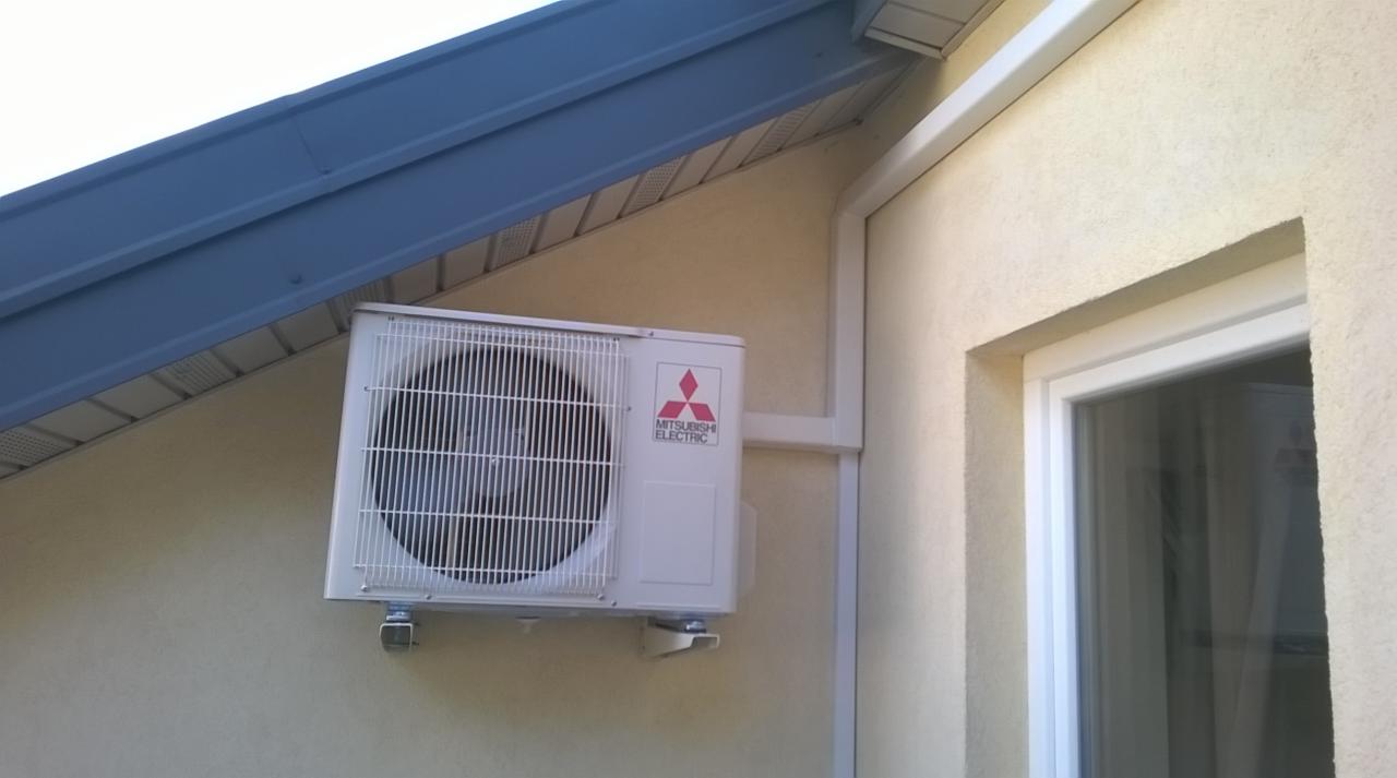 Biała jednostka zewnętrzna klimatyzacji Mitsubishi Electric zamontowana na kremowej ścianie budynku, widoczna pod niebieskim okapem dachowym i obok białej ramy okiennej.