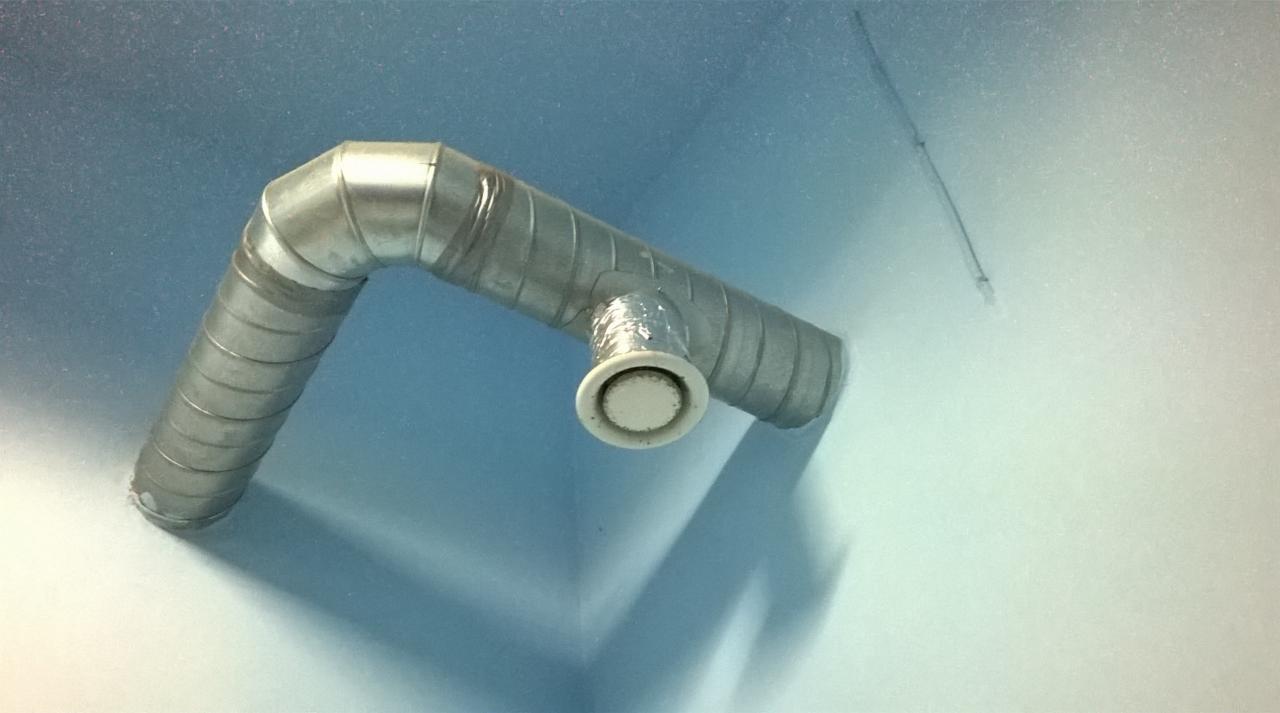 Metalowy kanał wentylacyjny z okrągłym wylotem na suficie w rogu pomieszczenia, widok z dołu.