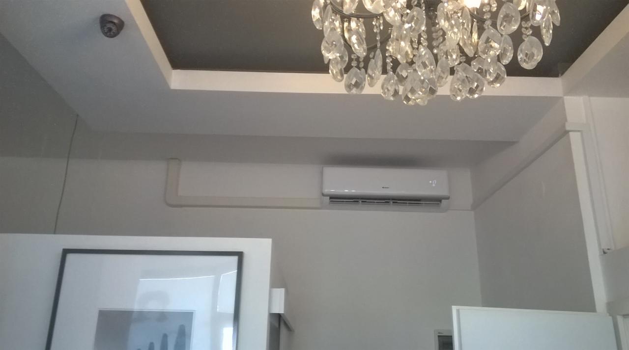 Biała jednostka klimatyzacyjna zamontowana na ścianie w pomieszczeniu z kryształowym żyrandolem i obrazem w czarnej ramie.