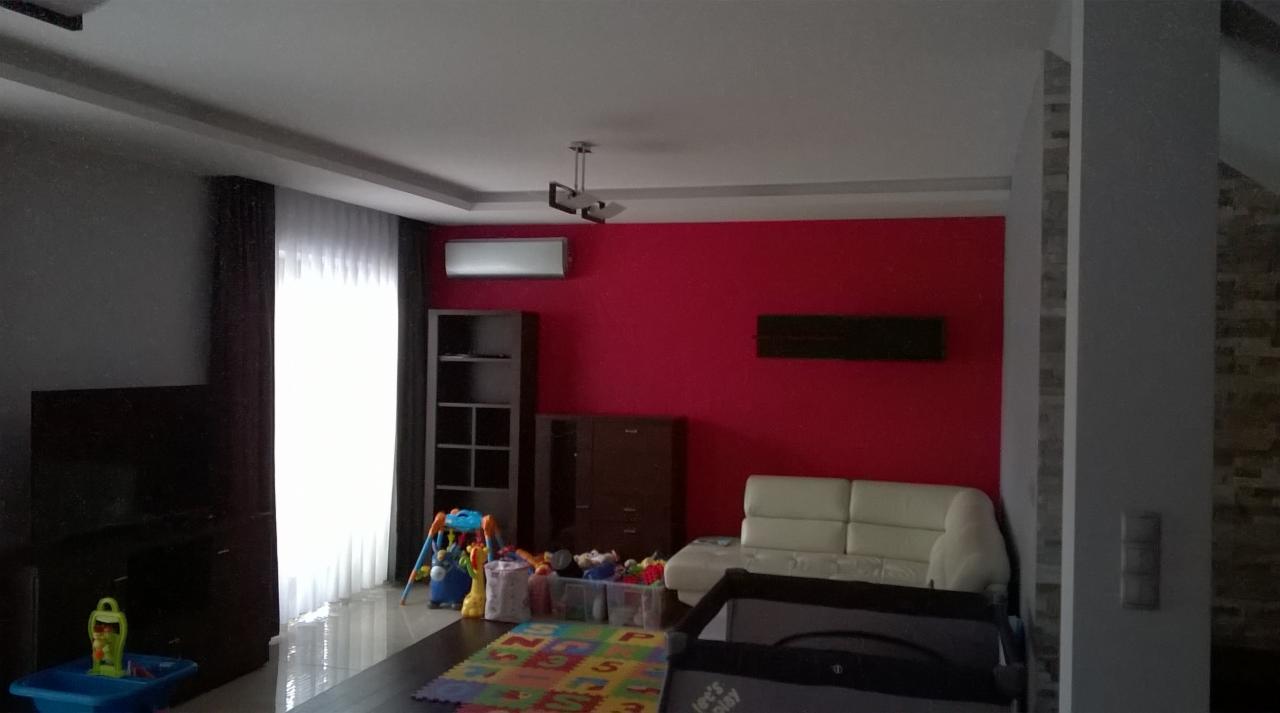 Klimatyzator ścienny zamontowany w pokoju dziecięcym z czerwoną ścianą, widoczny również dywan z literami, biała sofa i zabawki.